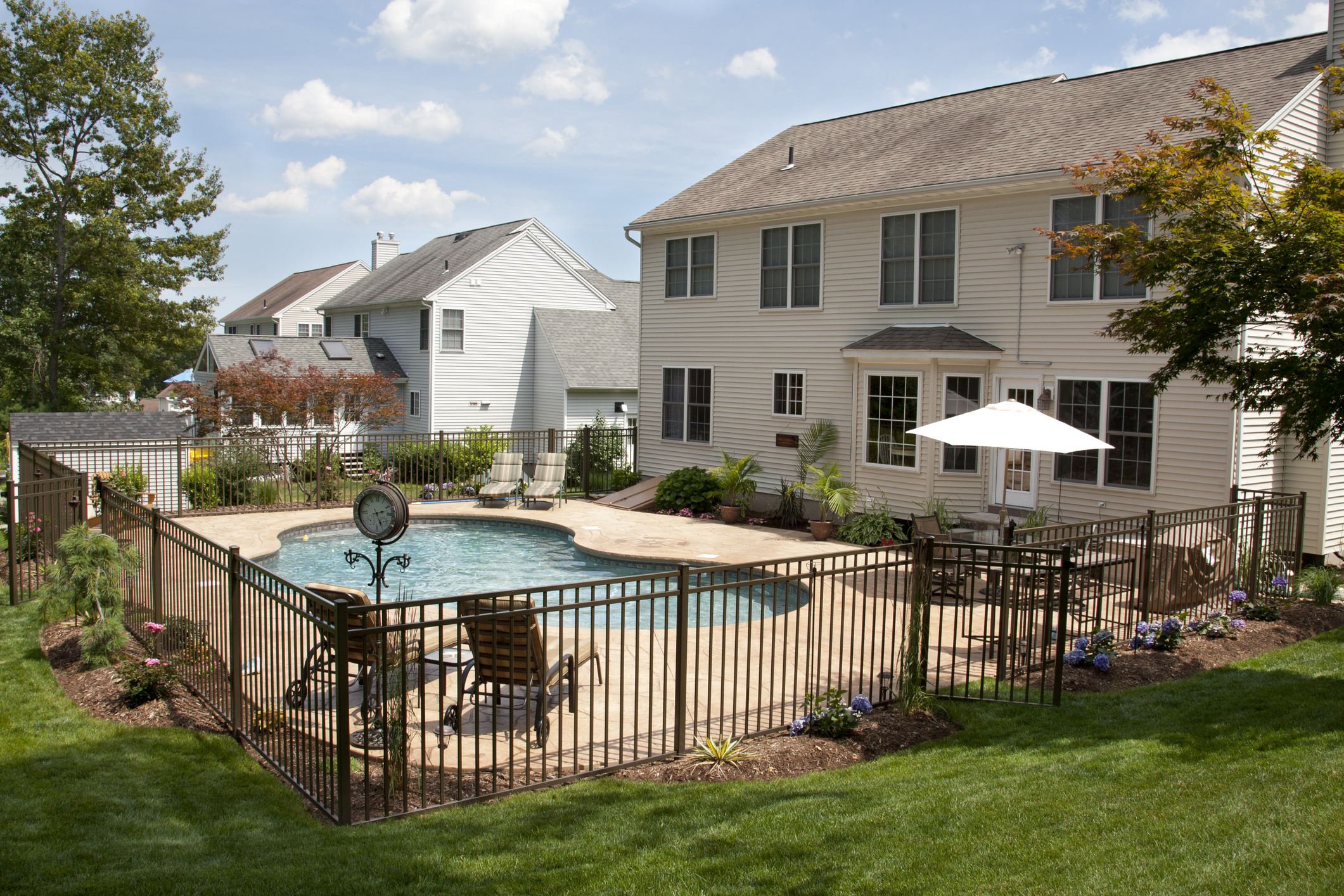 Lush, resort-like backyard salt water swimming pool with flagstone stamped concrete patio behind colonial style home.  Bronze fencing is seen around the pool area of this suburban backyard.