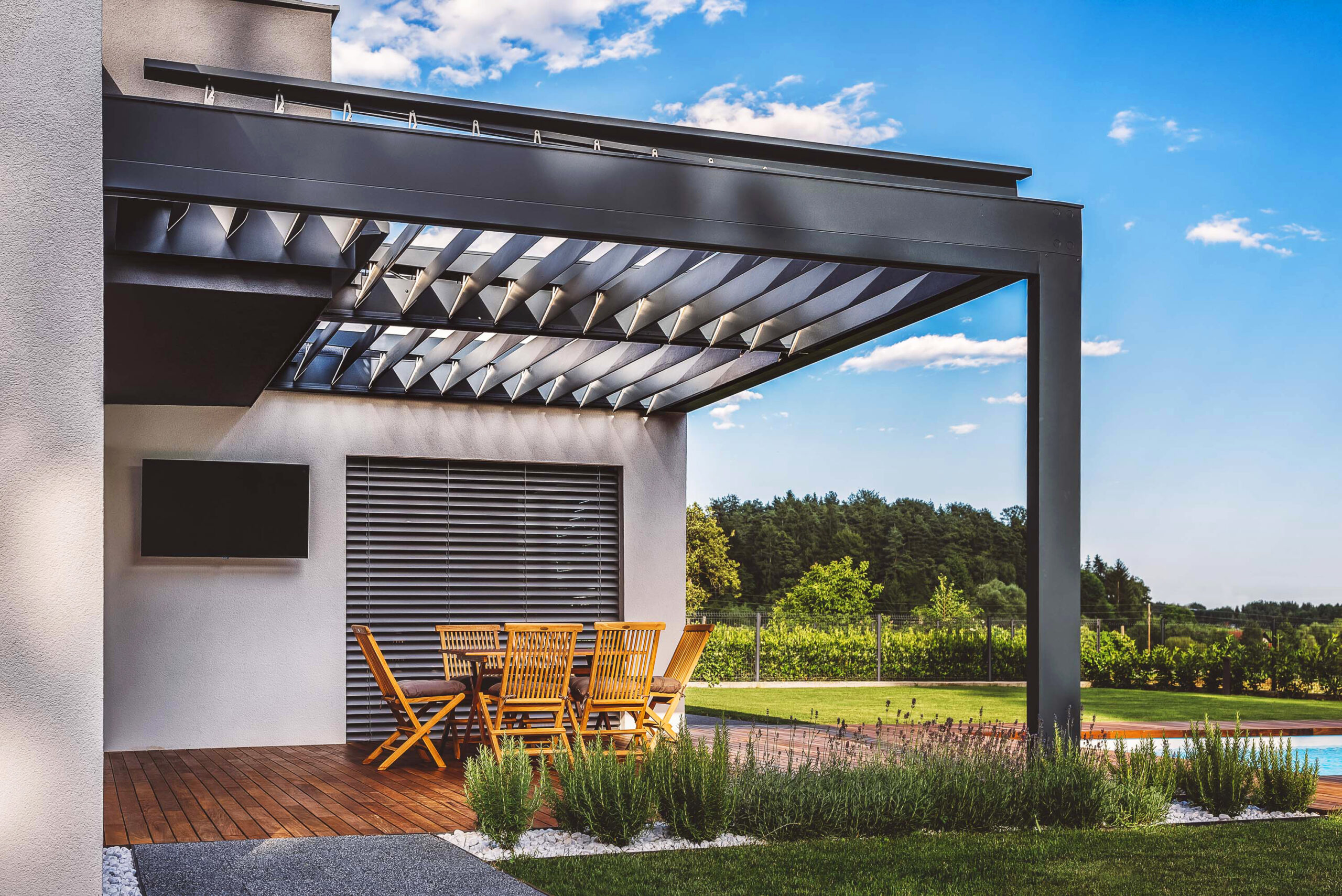 A modern outdoor patio with a pergola, featuring wooden furniture and a swimming pool in the background. The setting is surrounded by greenery and a clear blue sky.