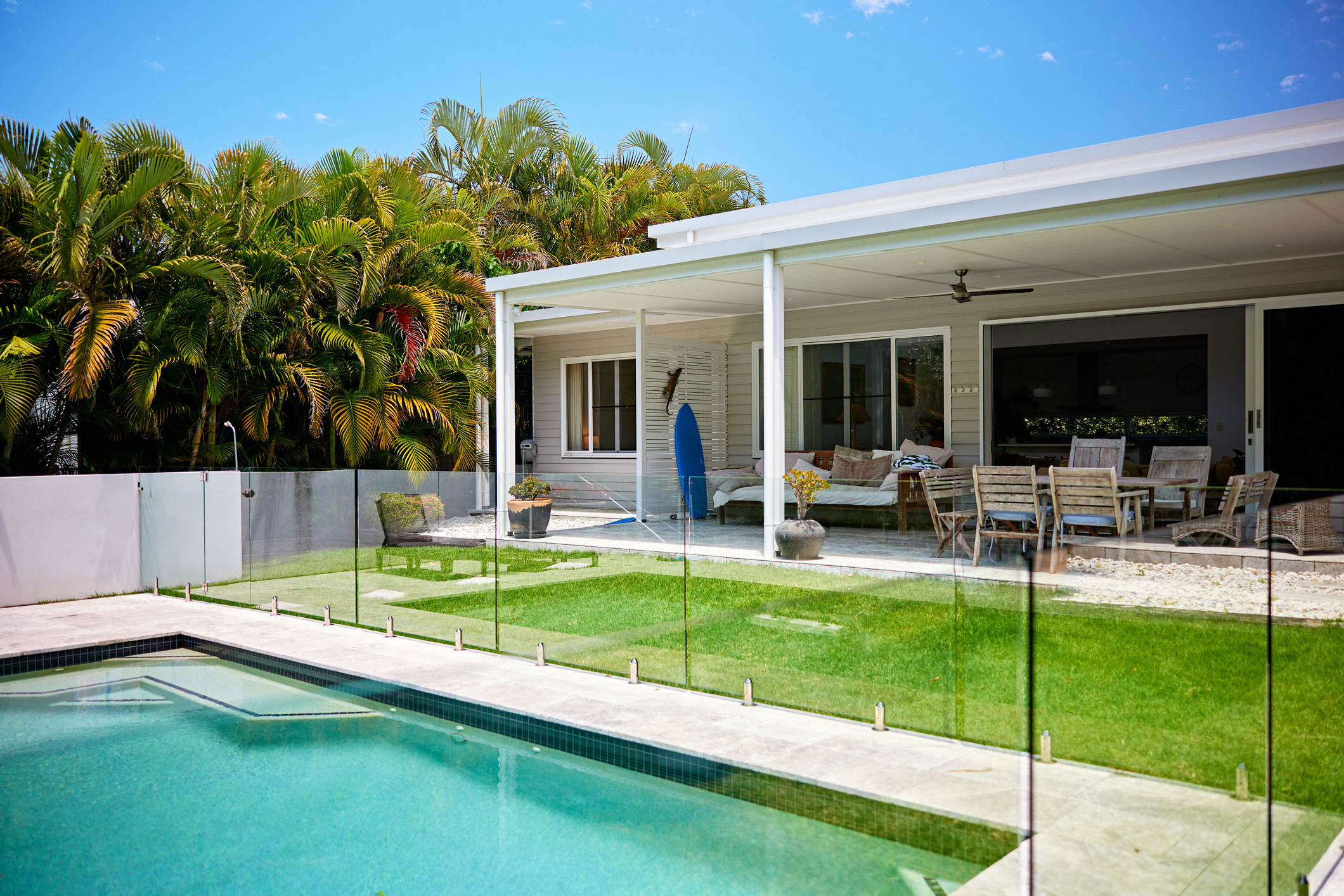 View across glass-enclosed swimming pool surrounded by green lawn, covered patio with dining table, chairs, and sofa, sliding door access to home interior.
