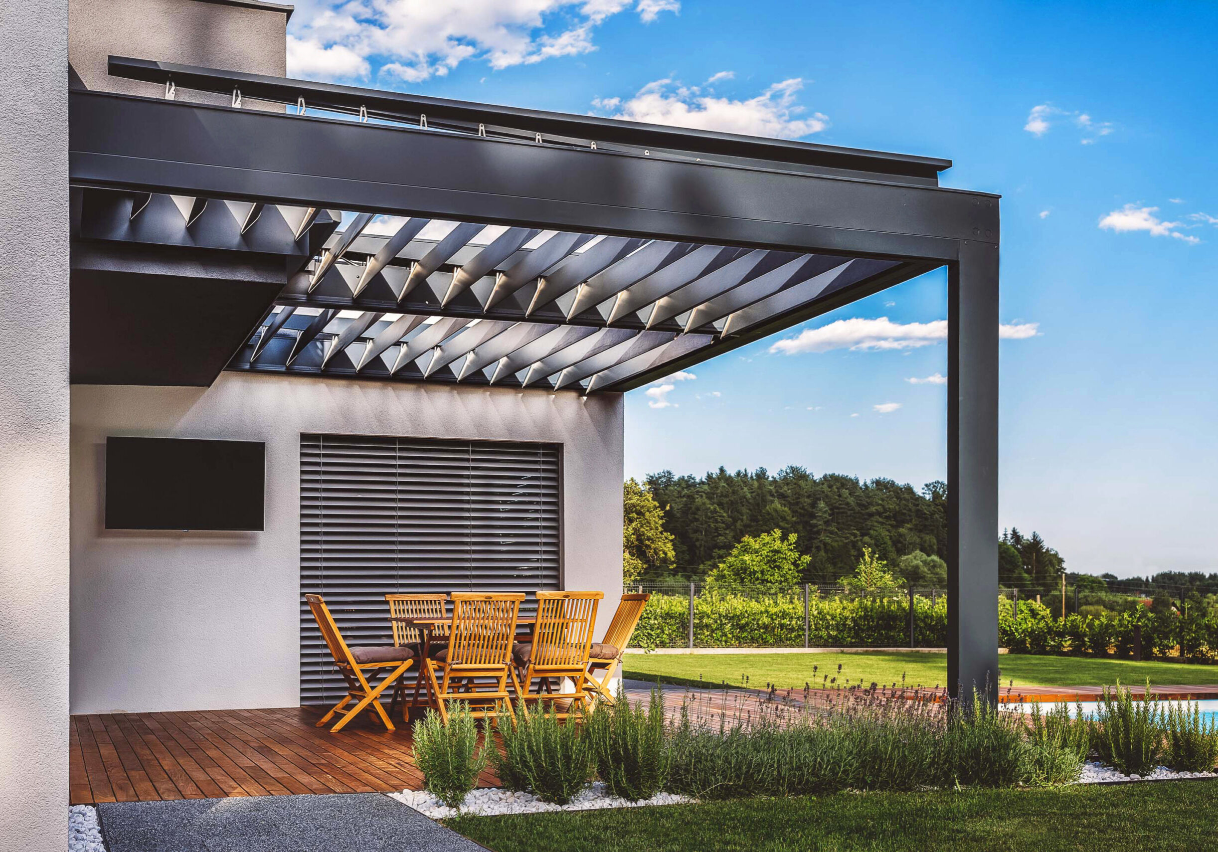 A modern outdoor patio with a pergola, featuring wooden furniture and a swimming pool in the background. The setting is surrounded by greenery and a clear blue sky.